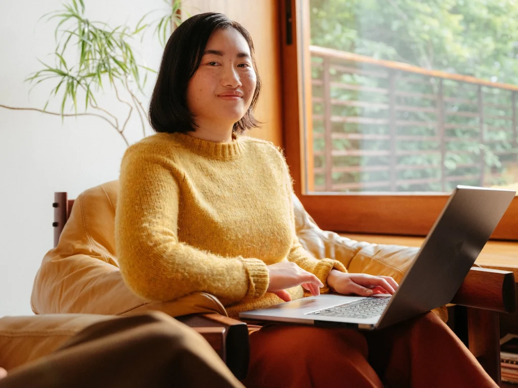 Kvinna i gul jumper som sitter på en soffa och ler mot kameran och har en laptop på knät